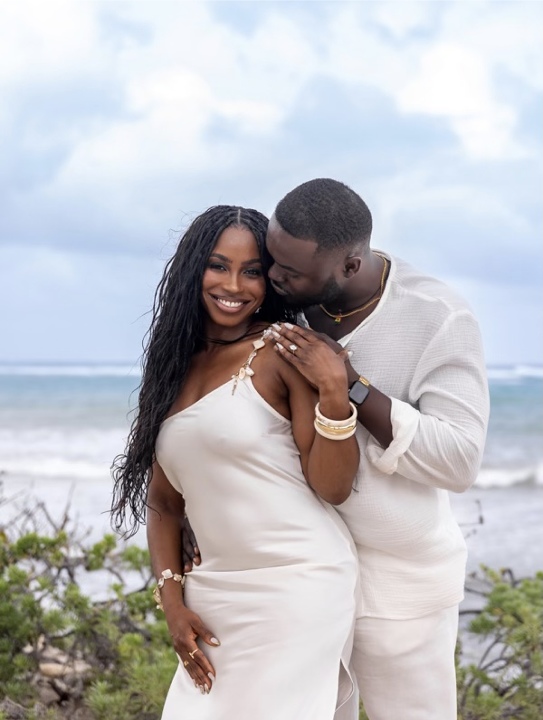 An engaged couple stands on a beach with the ocean in the background. The woman is smiling and wearing a white dress, and is wearing an engagement ring on her left hand, while the man, dressed in a light-colored shirt, stands behind her and kisses her on the temple. They are both adorned with bracelets and look joyful.